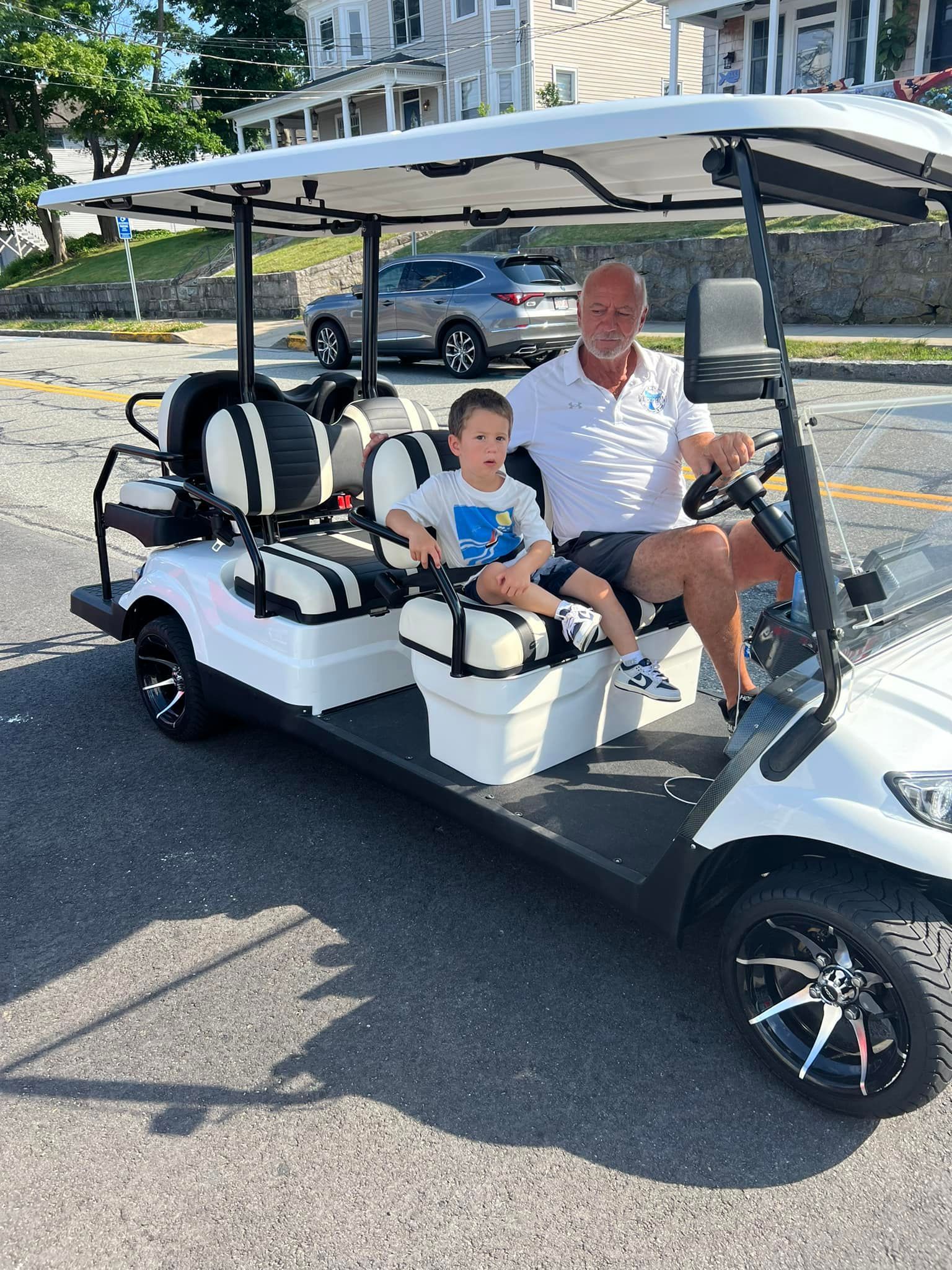 Man and child in a white golf cart on a street, man driving, child sitting in front, sunny day.