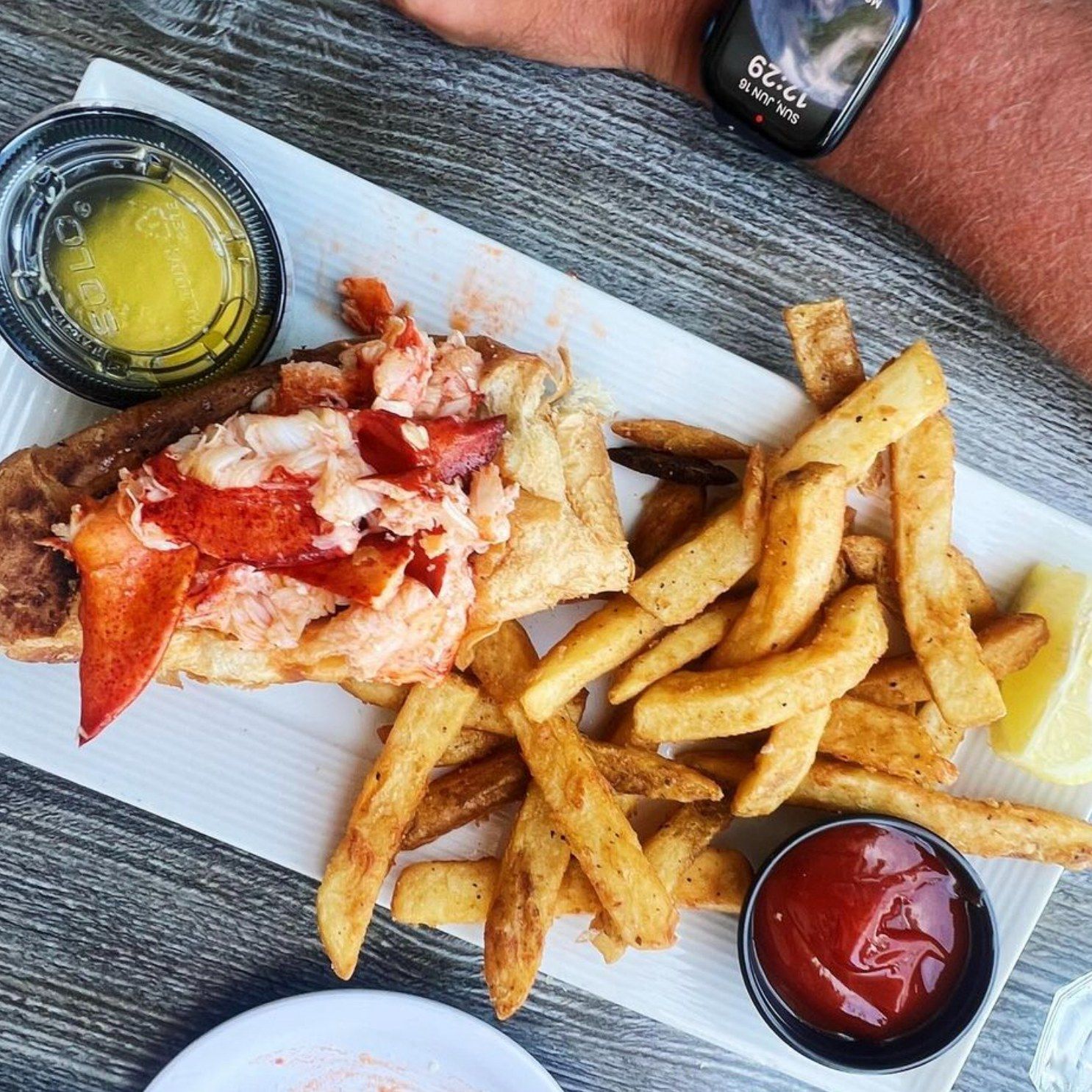 Lobster roll with fries, butter, and ketchup on a white rectangular plate.