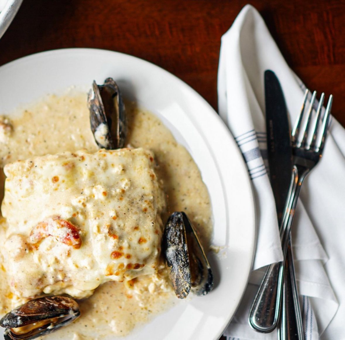 Plate of fish with creamy sauce and mussels, next to a napkin, knife, and fork.