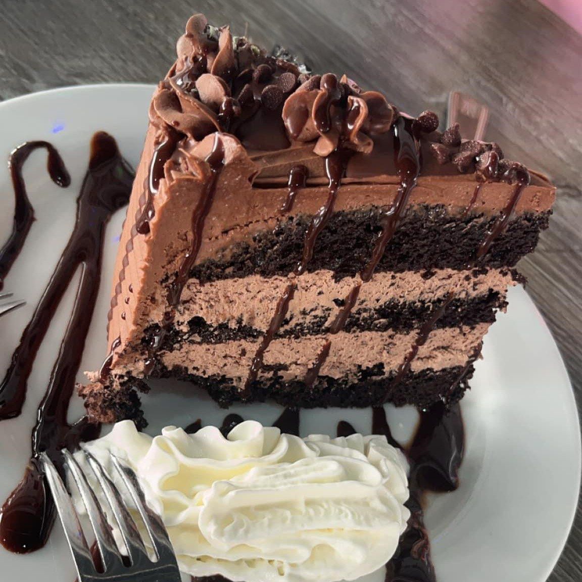 Slice of layered chocolate cake with frosting, drizzled chocolate sauce, whipped cream on plate.