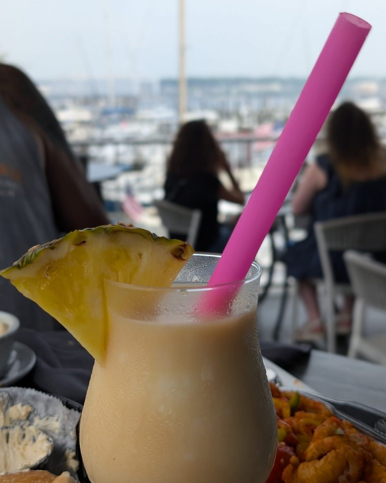 Pina colada cocktail with pink straw and pineapple garnish, set on a table with a blurred background of a waterfront.