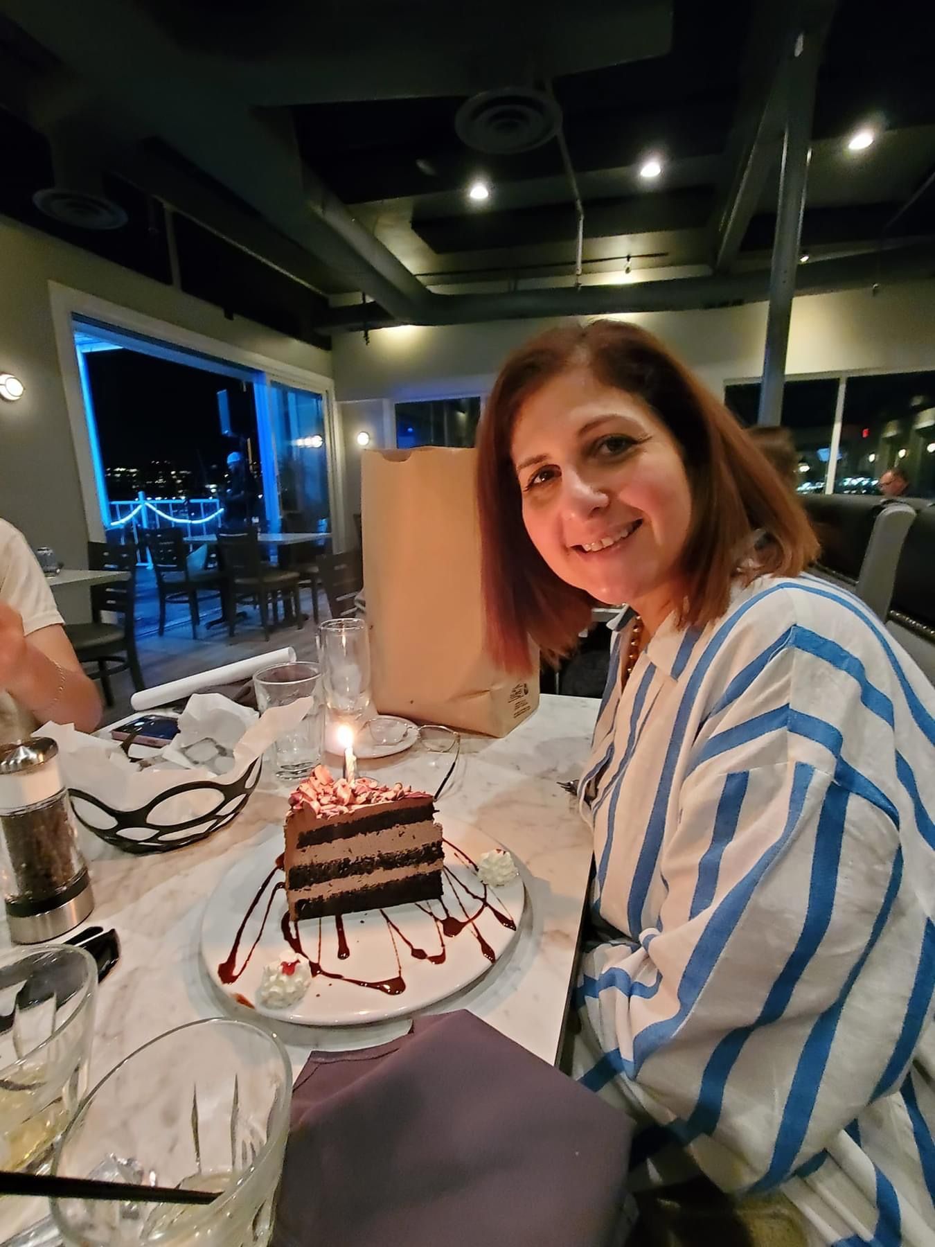 Woman at a restaurant smiles, birthday cake with candle, striped shirt.