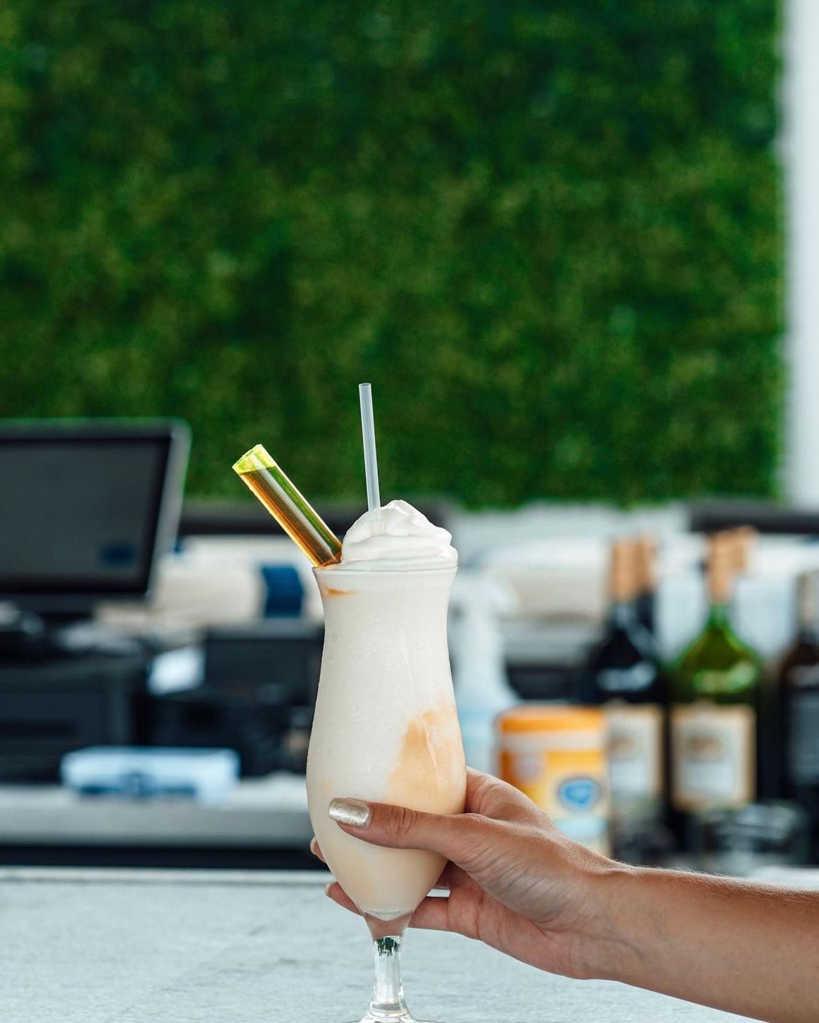 Hand holding a frosted cocktail with whipped cream, yellow garnish, and straw, set against a green wall.