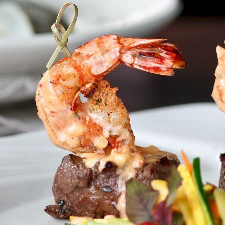 Grilled shrimp atop seared steak, skewered with a decorative pick, served on a white plate.