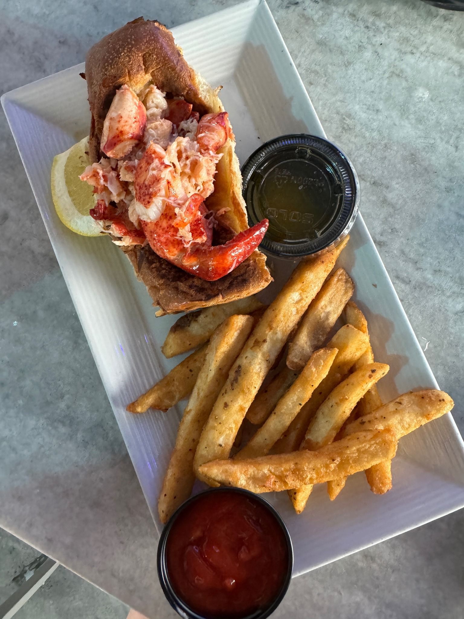 Lobster roll, french fries, lemon, and dipping sauces on a white rectangular plate.