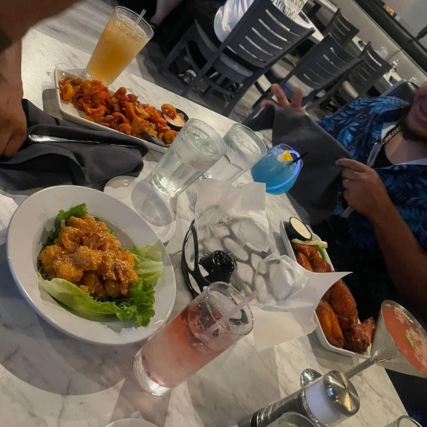 Table with drinks, appetizers, and people at a restaurant. Dishes include fried shrimp, wings, and drinks.