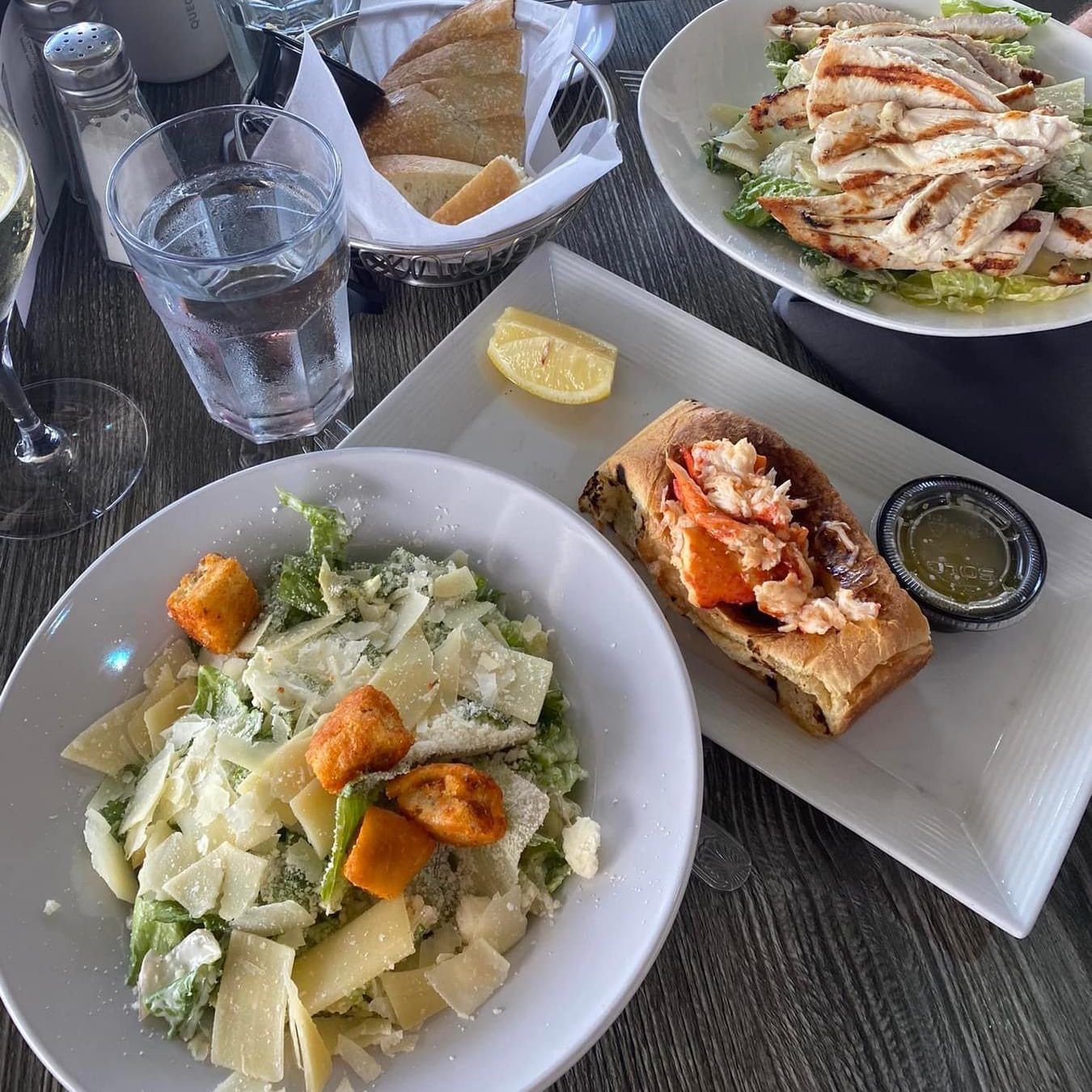 A table set with Caesar salads, a lobster roll, bread, water, and grilled chicken salad.