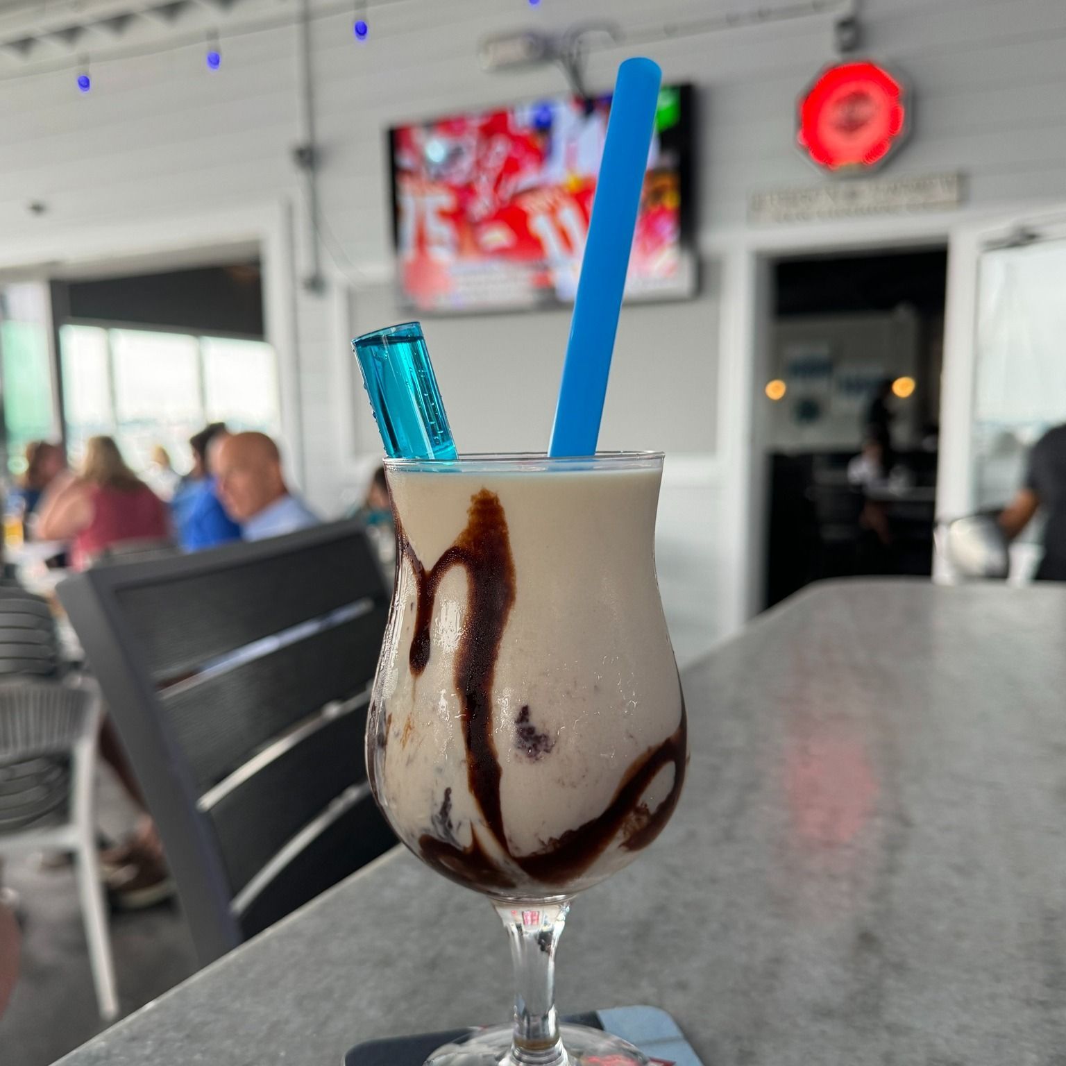 Creamy drink in a glass with chocolate drizzle, blue straw, and a blue ice pack garnish on a bar.