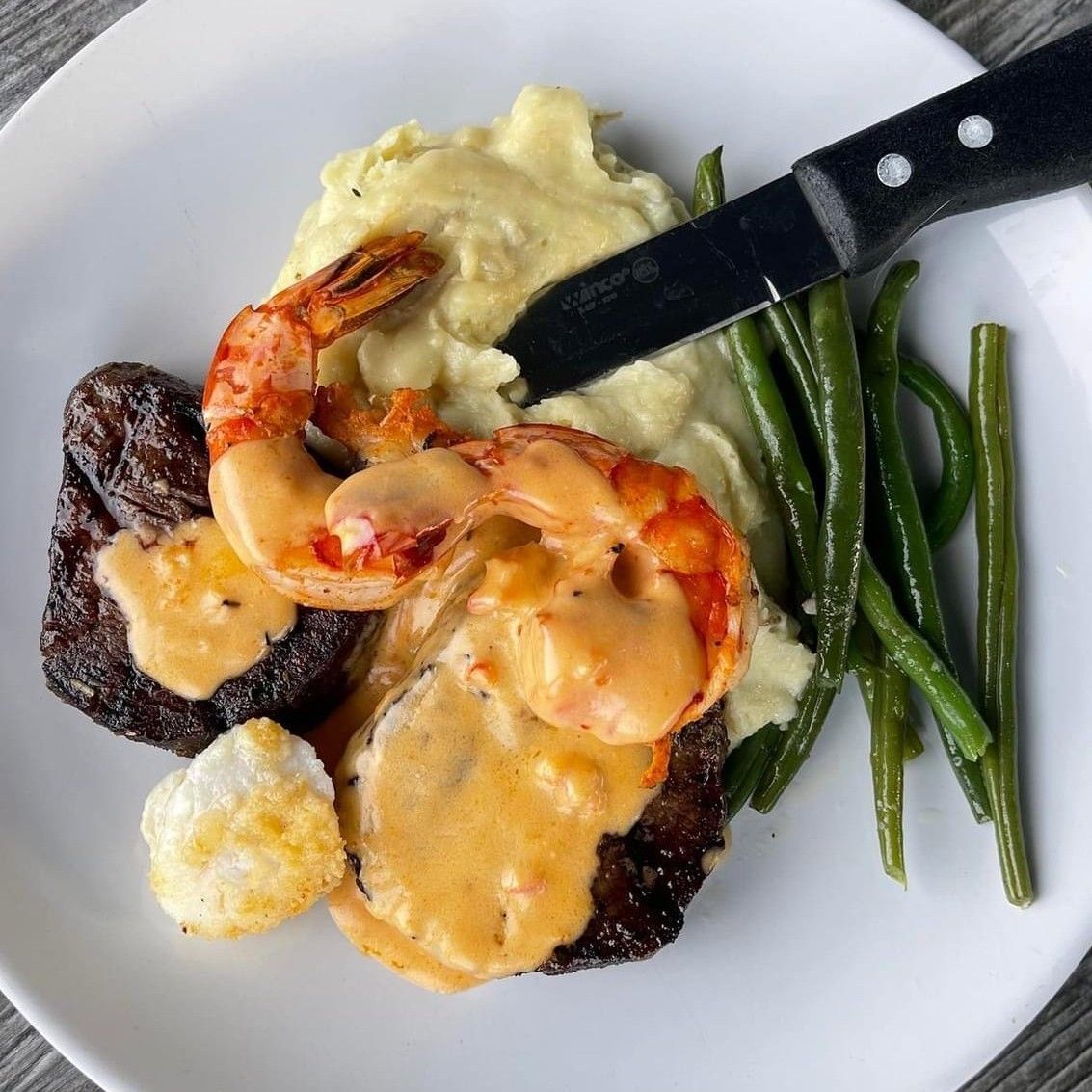 Steak, shrimp, mashed potatoes, and green beans on a white plate with a knife.
