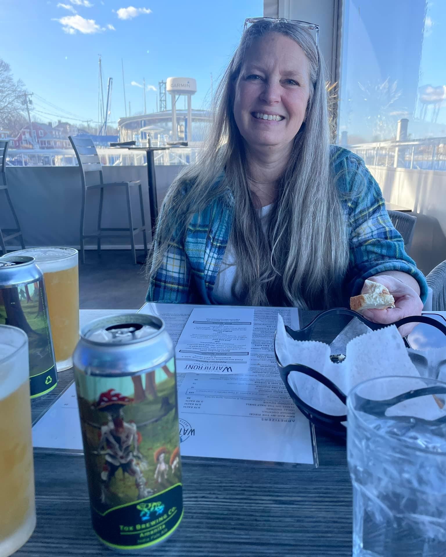 Woman smiling, sitting at a table with beer and a menu. Outdoors, sunny day.