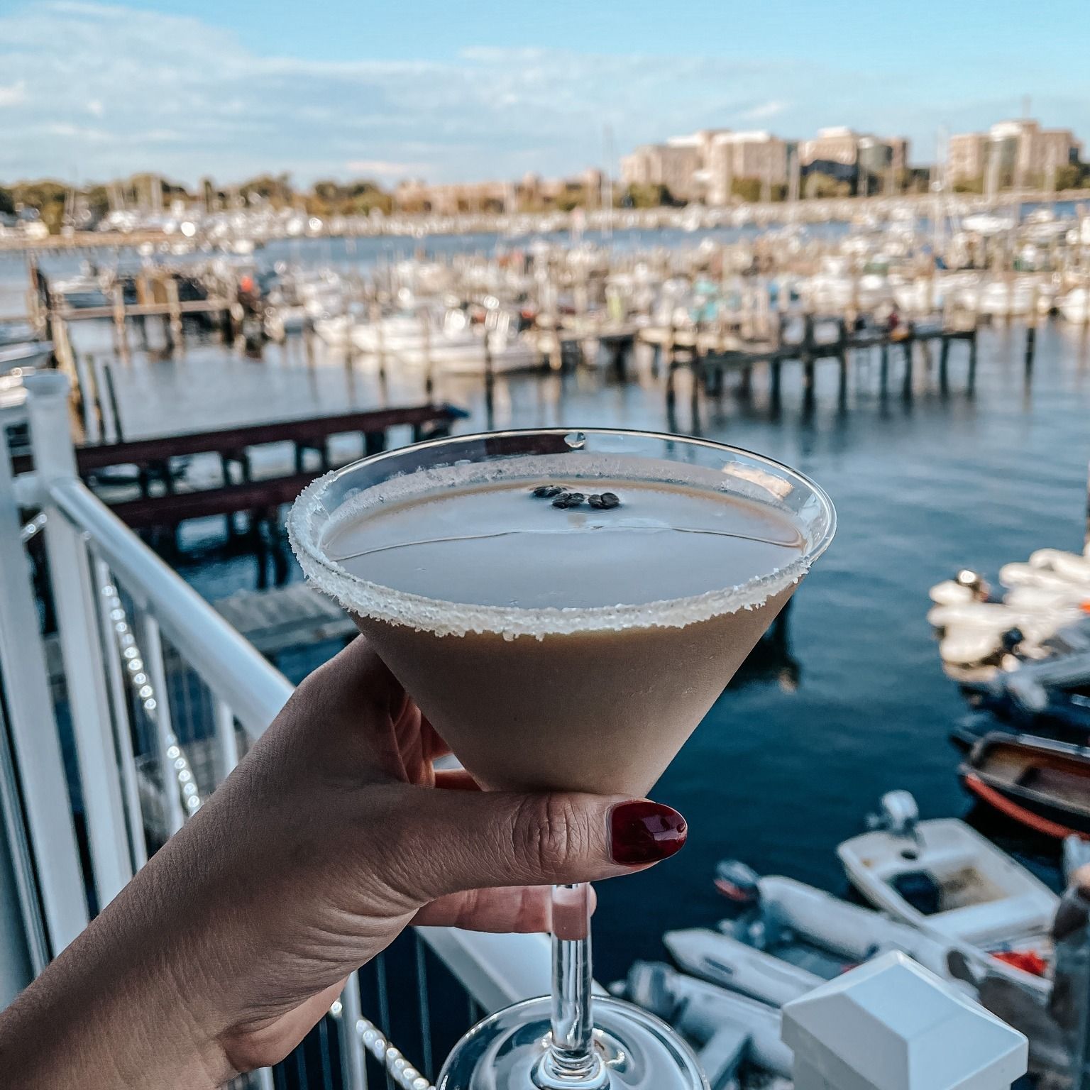 Hand holding a martini glass with brown drink and coffee beans, overlooking a marina.