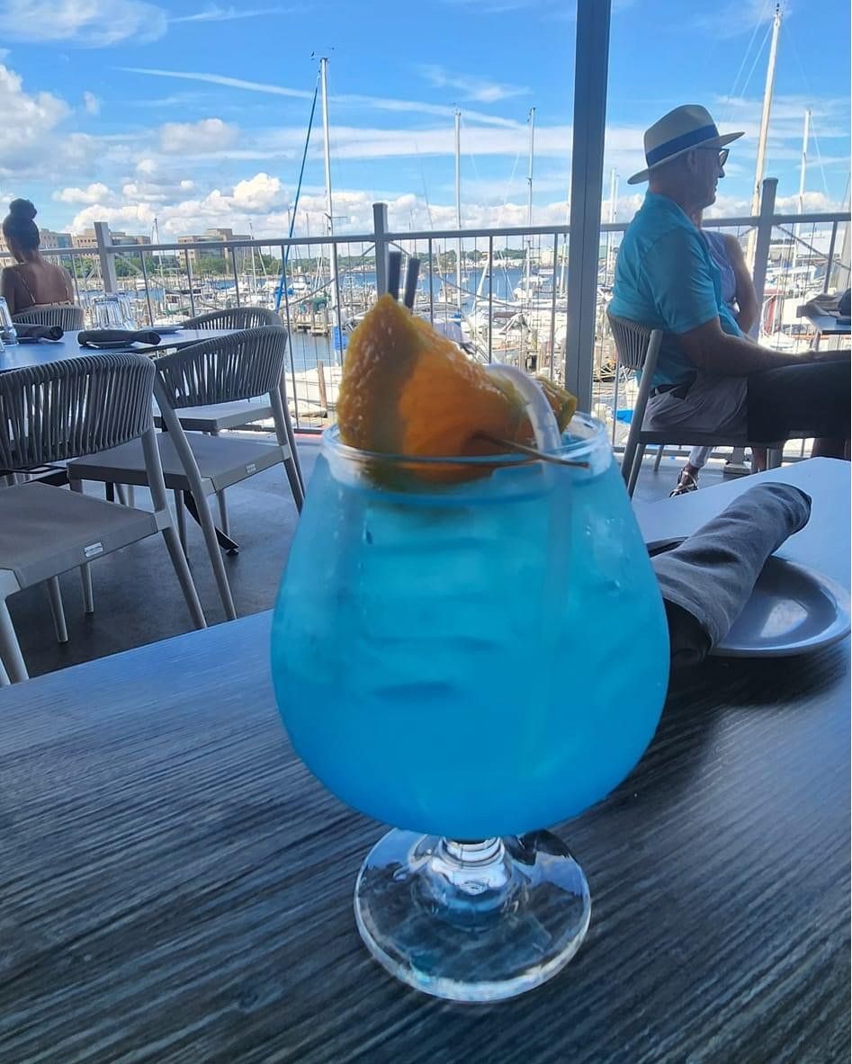 Blue cocktail with orange slice garnish on a table overlooking a marina.