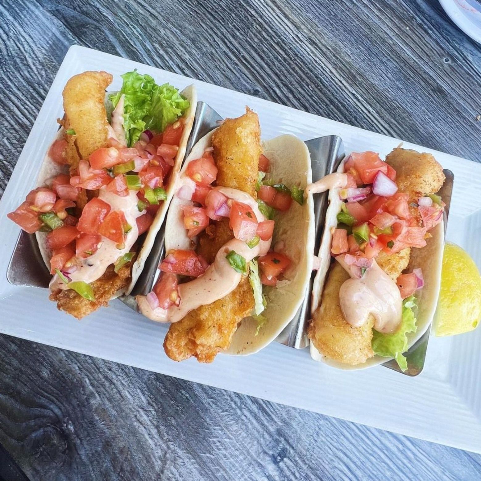 Three fish tacos with salsa and sauce on a white plate with a lemon.