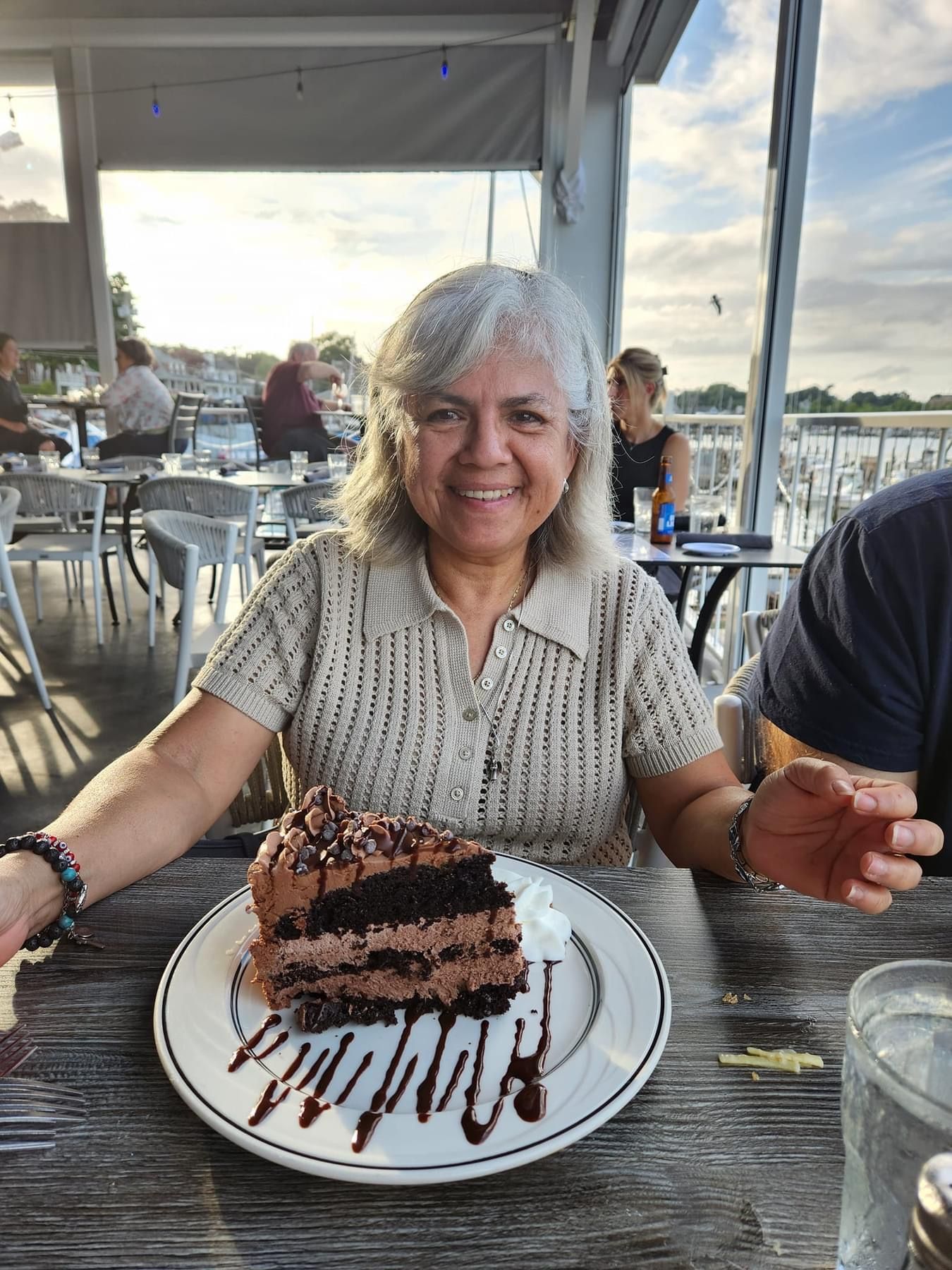Woman smiling at camera, holding plate with chocolate cake. Outdoor restaurant setting.