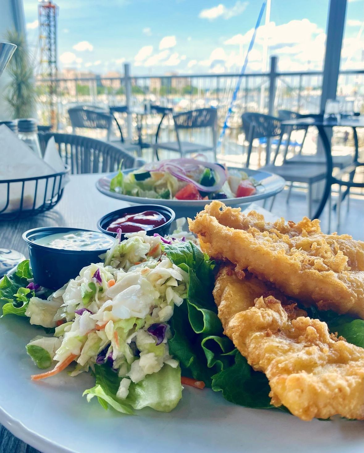 Plate of fried fish with coleslaw, dipping sauces, and a salad on an outdoor patio overlooking a marina.