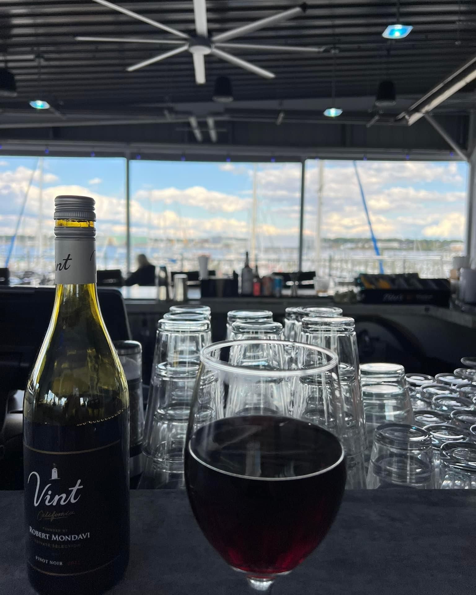Bottle and glass of red wine on a bar, overlooking a marina through large windows.