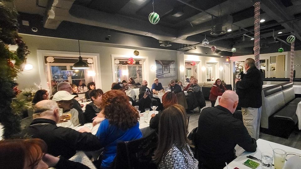 Restaurant interior with a person speaking to a seated audience at tables. Festive decorations are visible.