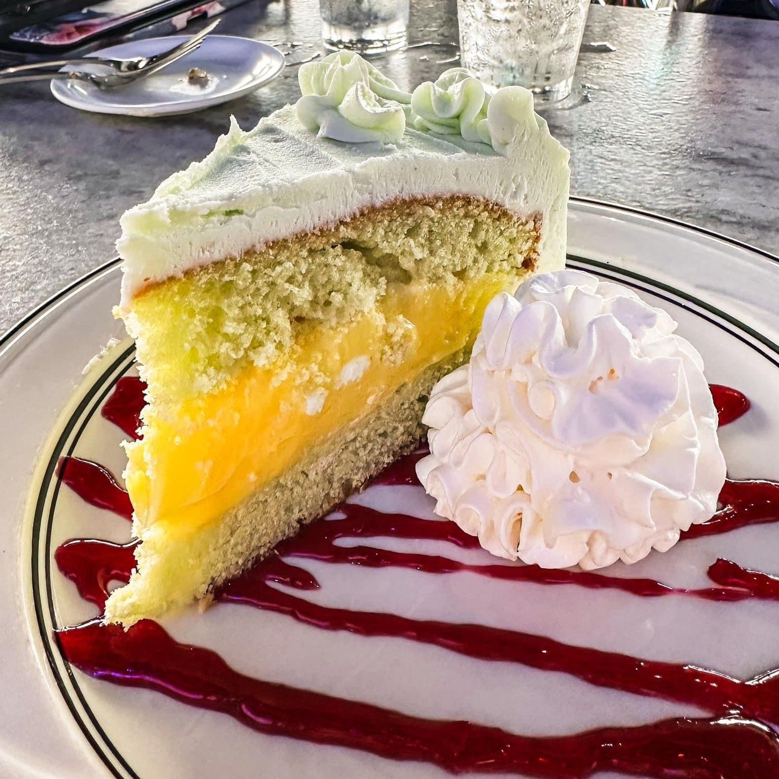 Slice of cake with layers of frosting, yellow filling, and whipped cream on a plate with red sauce.