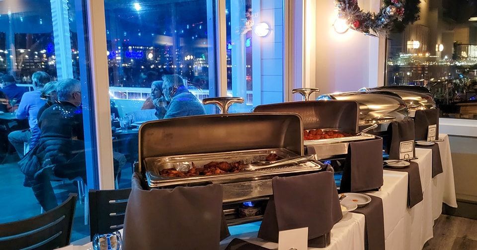 Buffet with covered food trays set up indoors near windows overlooking a city at night.