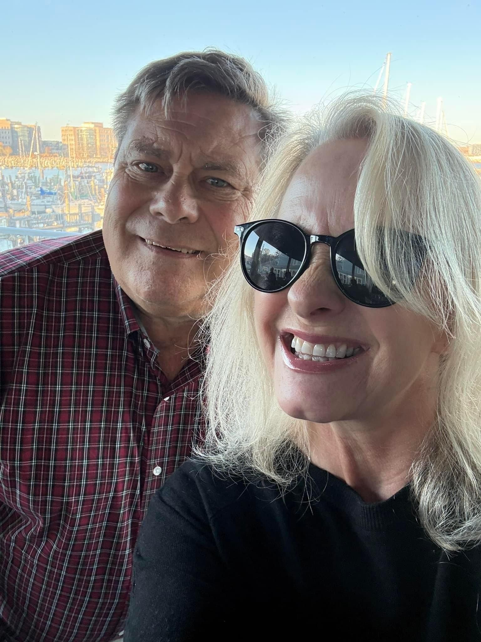 Man and woman smiling, taking a selfie outdoors with city and water background.
