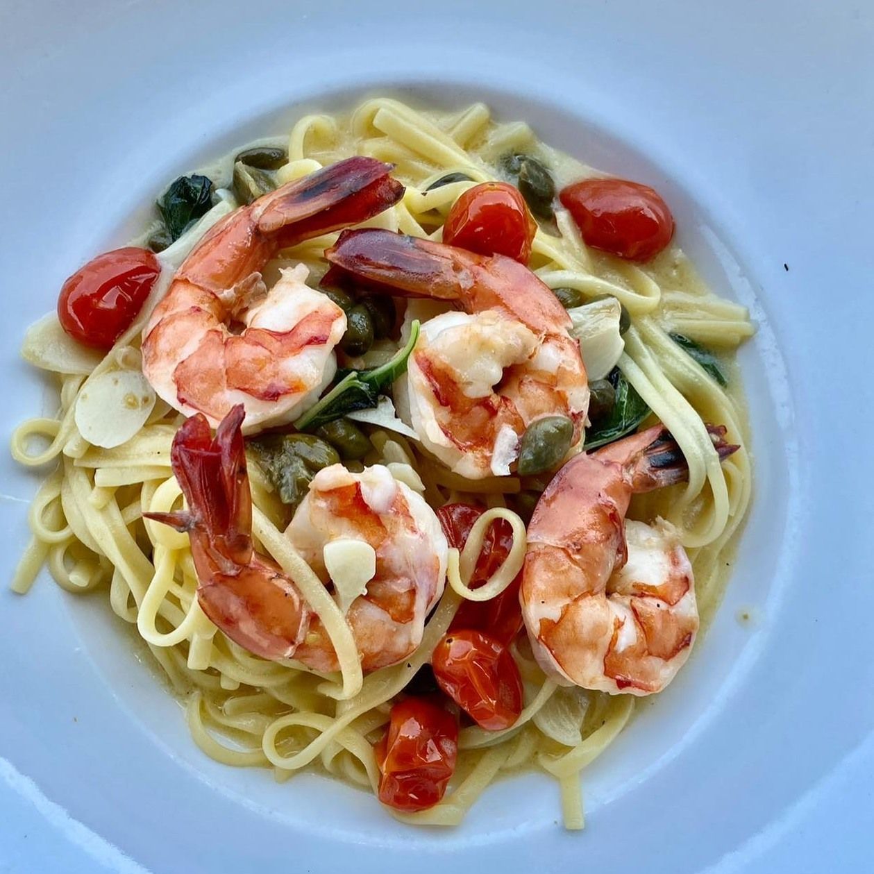 Linguine pasta with shrimp, cherry tomatoes, capers, and garlic in a white bowl.