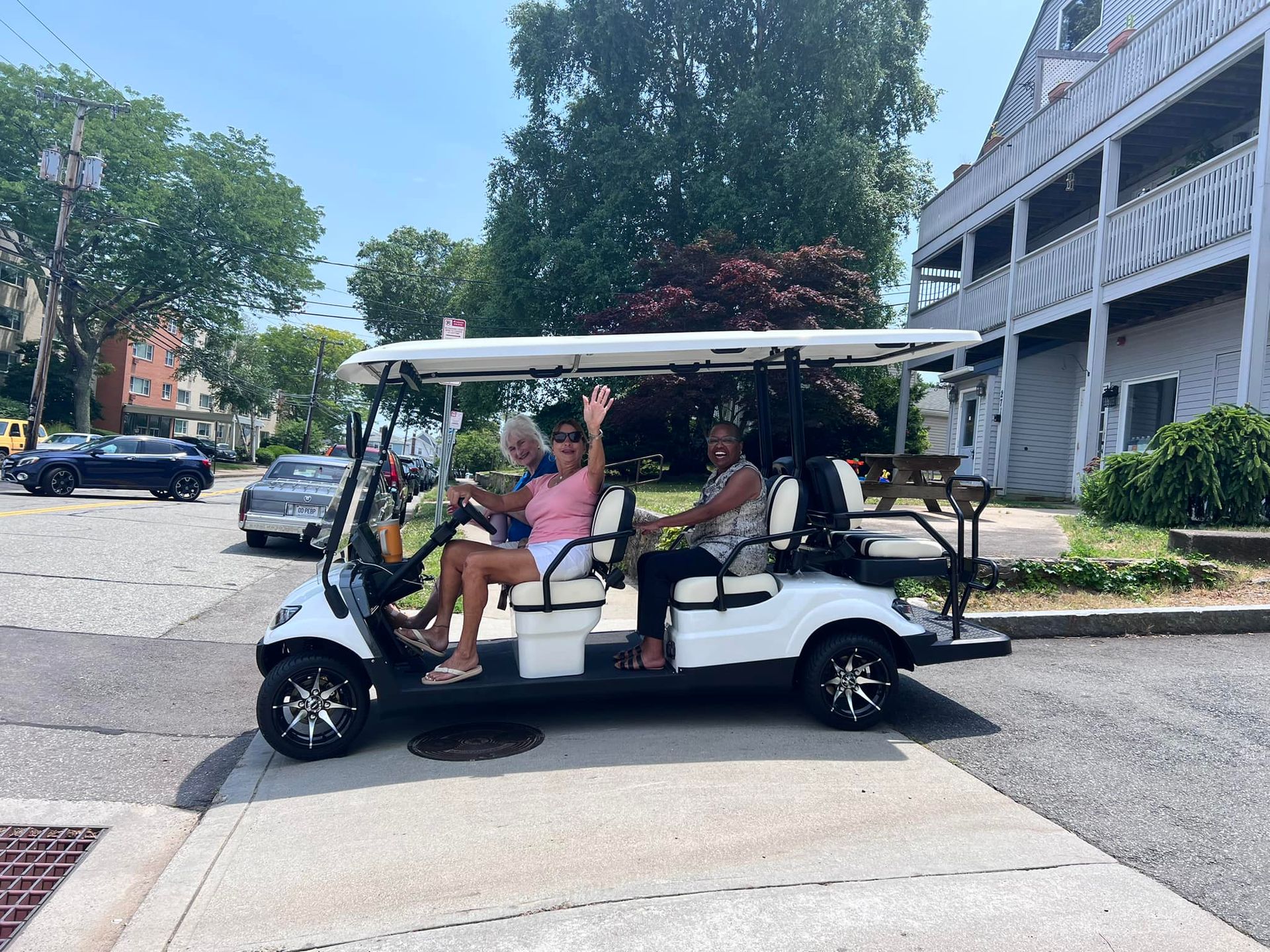 Three people wave from a white golf cart on a street, with buildings and cars visible in the background.