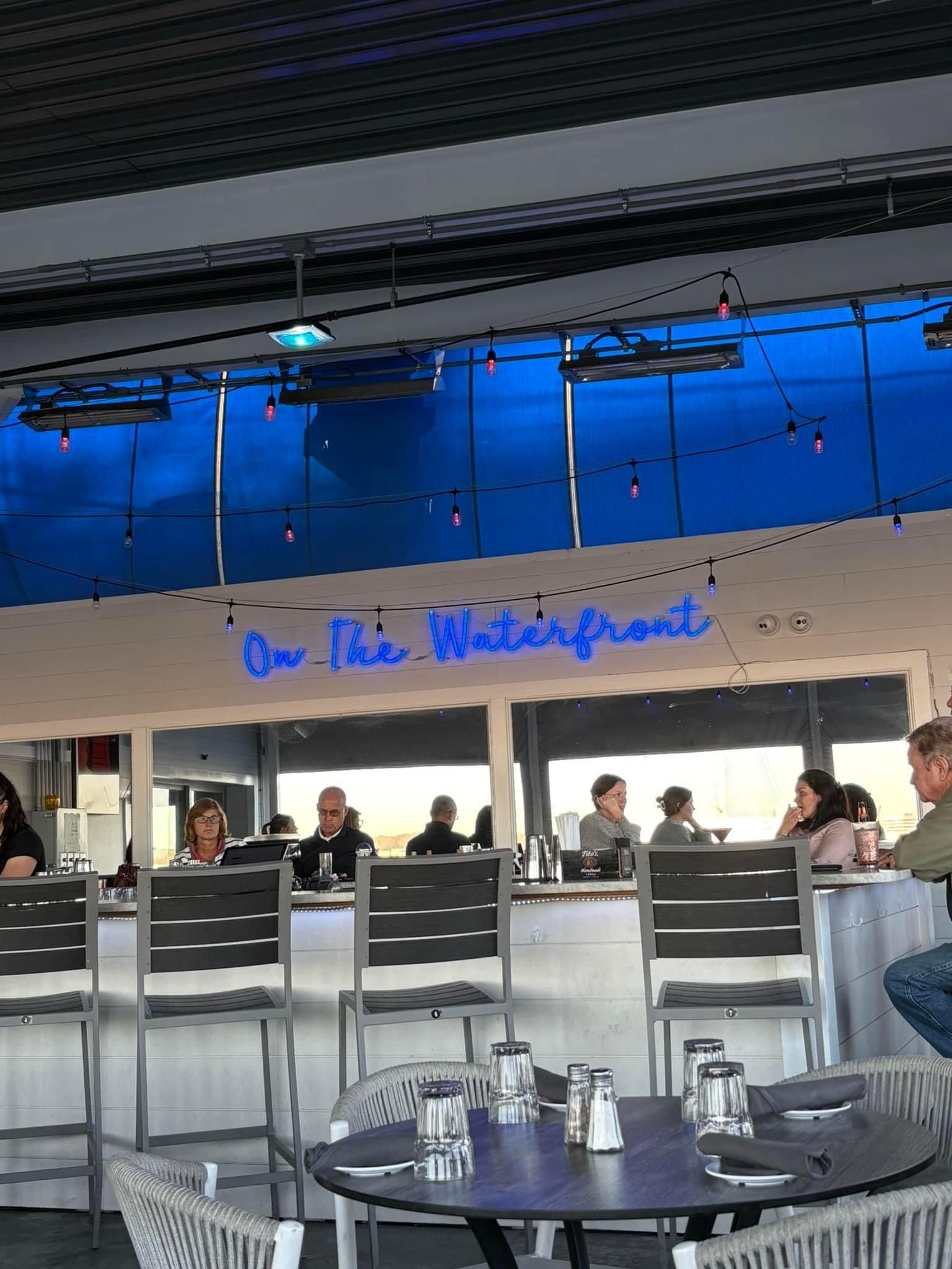 Restaurant with a bar and tables. People sit at the bar and tables under blue-lit windows.