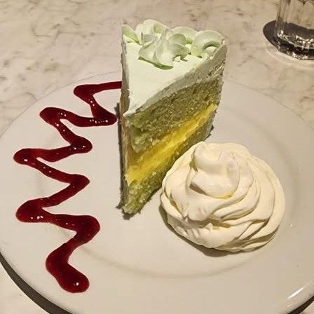 Slice of green cake with white frosting and yellow filling, whipped cream, and berry sauce on a white plate.