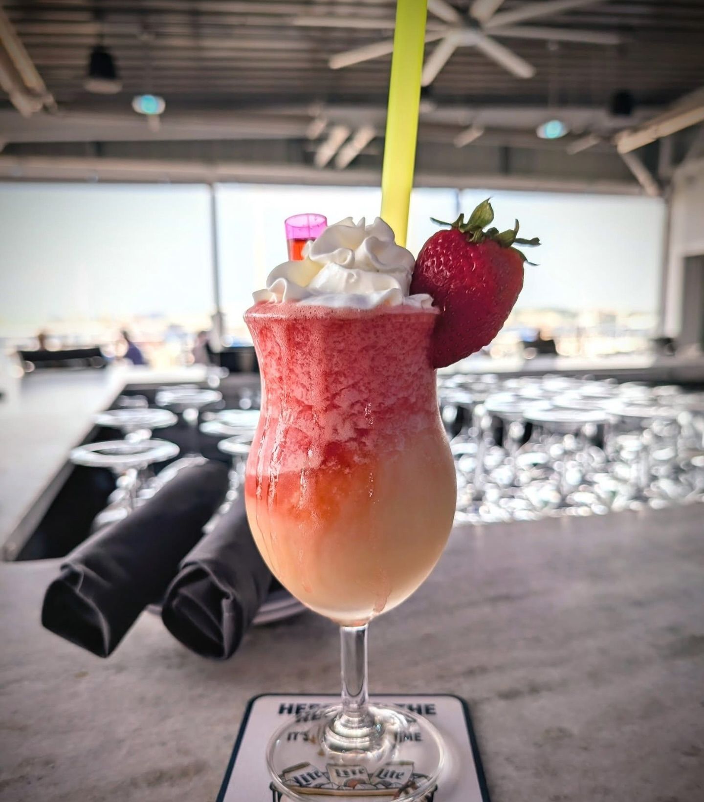 Strawberry daiquiri in a stemmed glass, topped with whipped cream and a strawberry, on a bar with a straw.