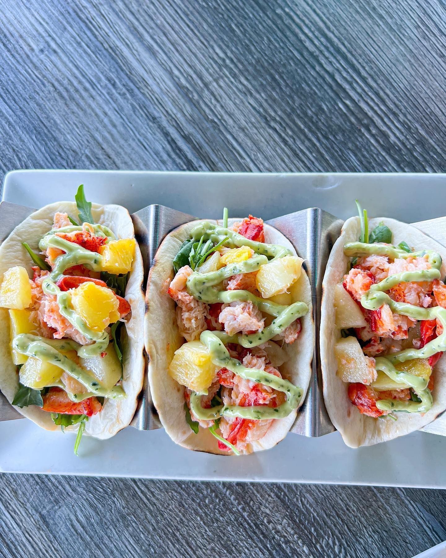 Three lobster tacos with pineapple and green sauce on a white plate.