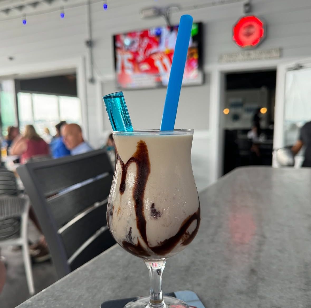 A cocktail in a stemmed glass with chocolate drizzle, blue straw, and a blue ice pop, set at a bar with patrons.