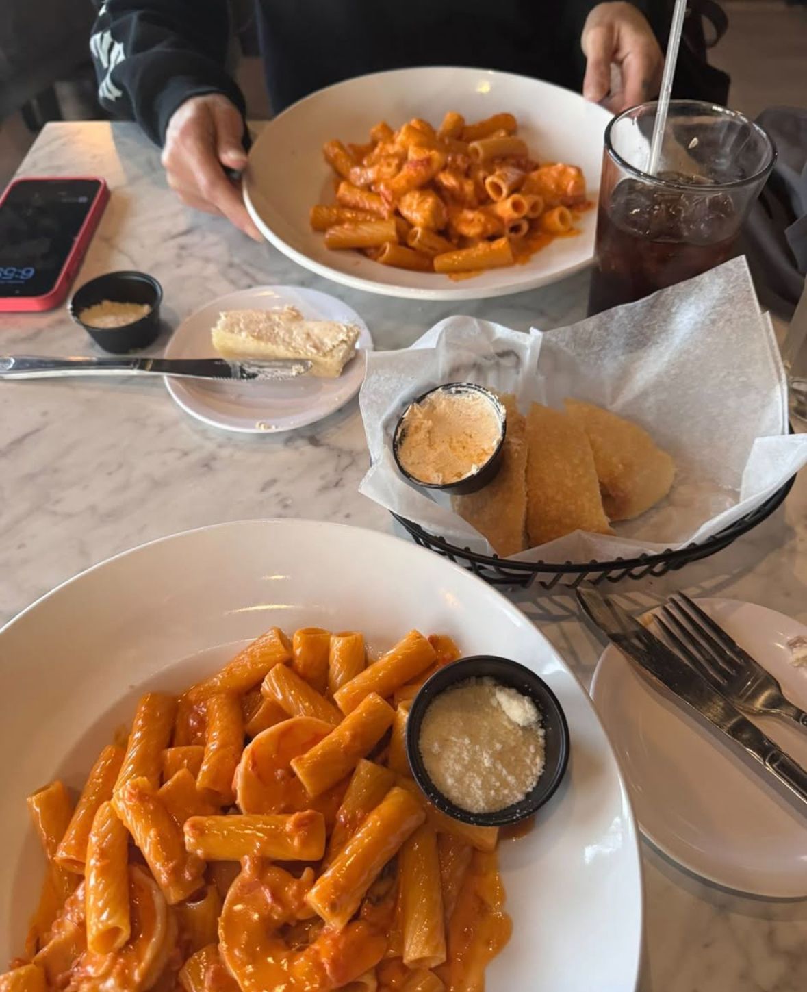 Two plates of pasta with orange sauce, bread, butter, and drinks on a marble table.