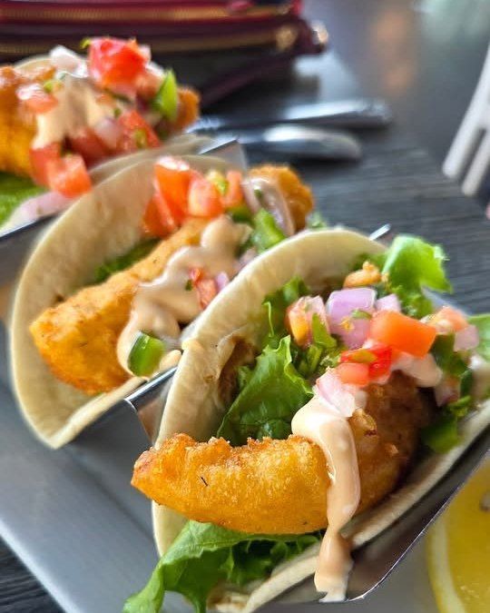 Three fish tacos with fried fish, salsa, and sauce on a gray plate.