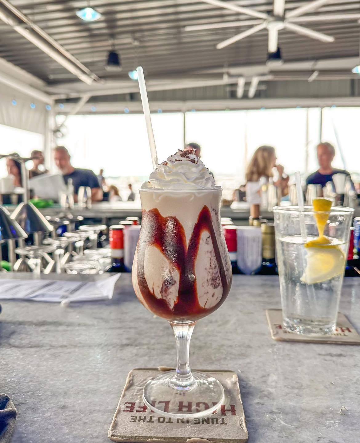 Tall sundae in a glass with dripping chocolate syrup, topped with whipped cream, on a bar.