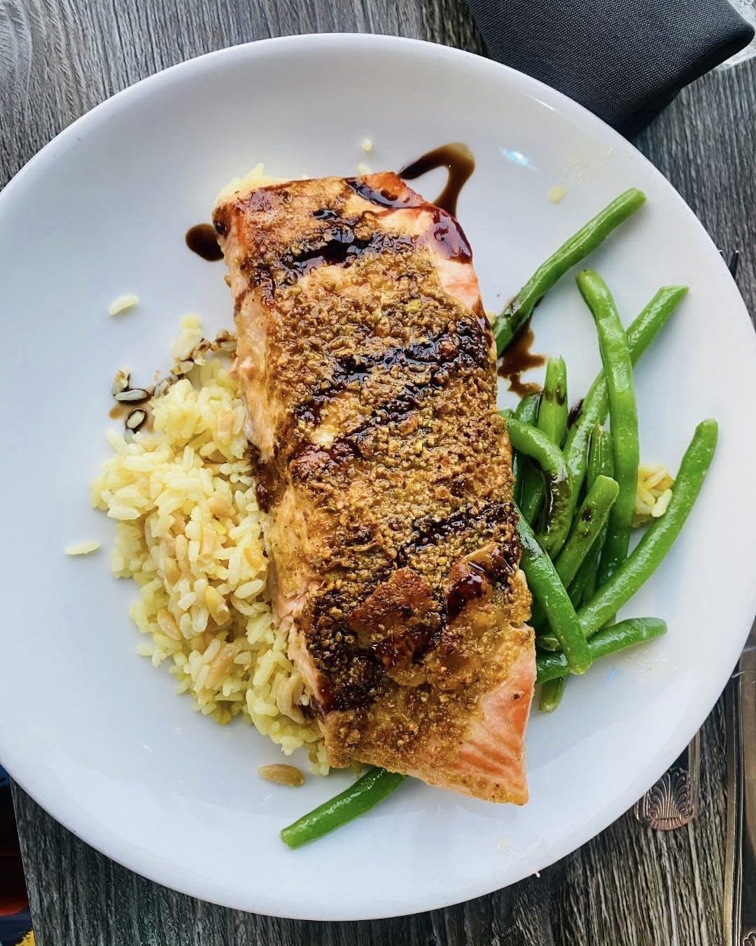 Salmon fillet with brown topping, rice, green beans, and balsamic glaze on a white plate.