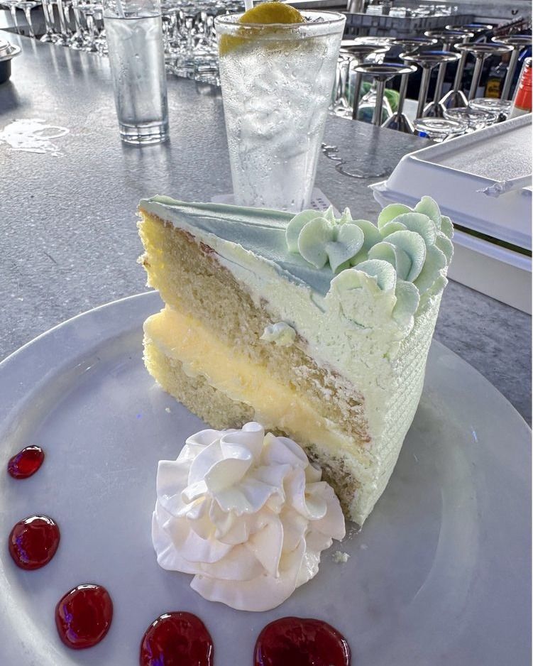 Slice of layered cake with white frosting, whipped cream, and berry sauce on a plate, next to a drink.