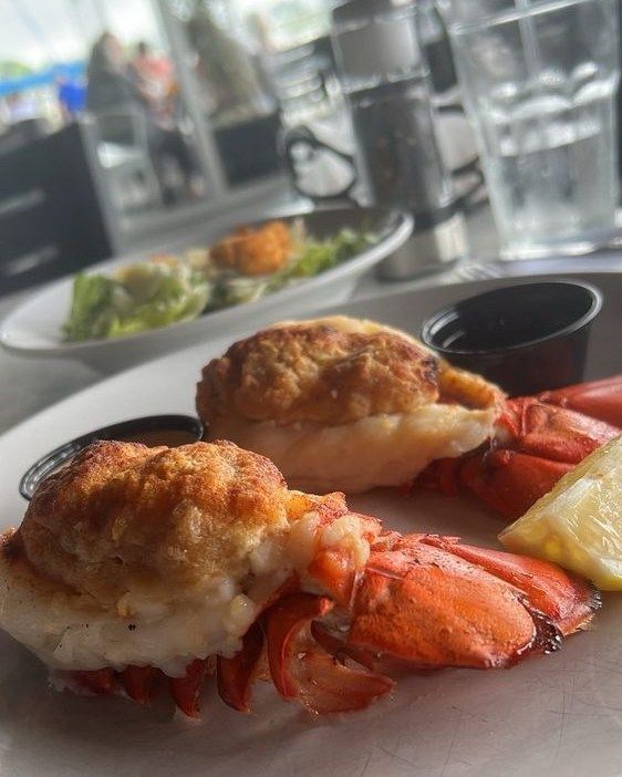 Lobster tails with a baked topping, served with sauce and lemon. A salad is in the background.