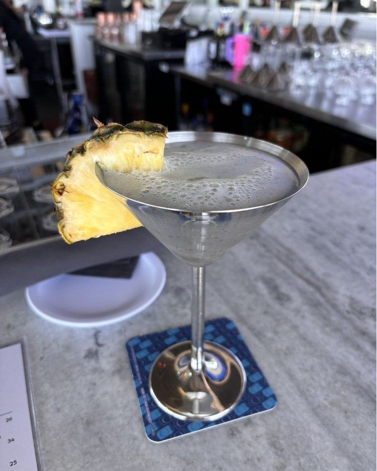 Cocktail in a silver glass, pineapple garnish, on a coaster at a bar.