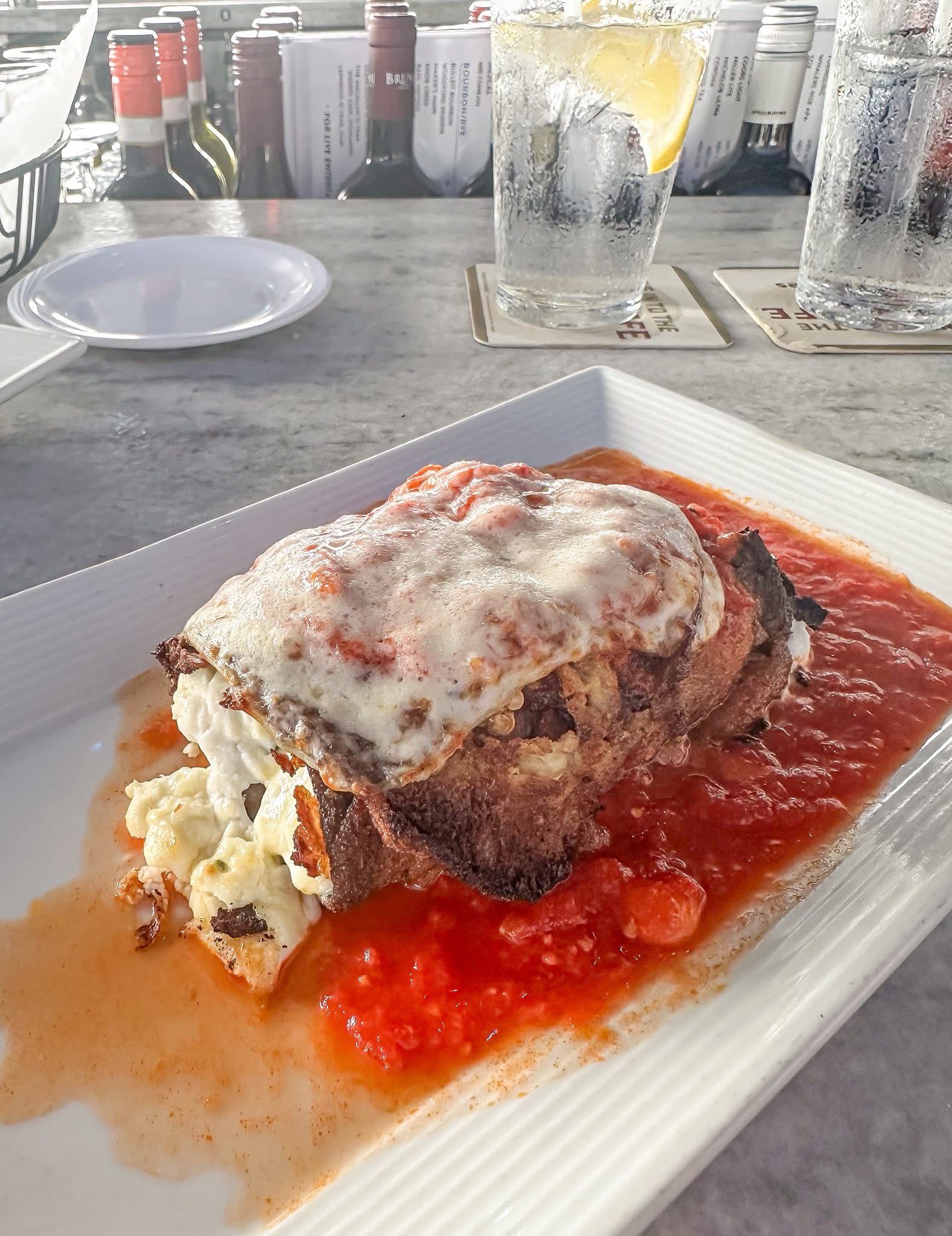 Lasagna on a white plate with tomato sauce, with drinks and wine bottles in the background.