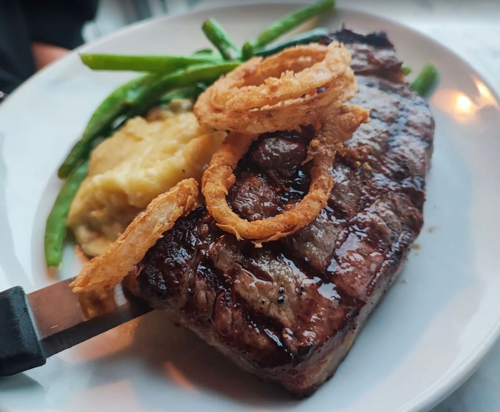 Steak dinner on a white plate with mashed potatoes, green beans, and onion rings.