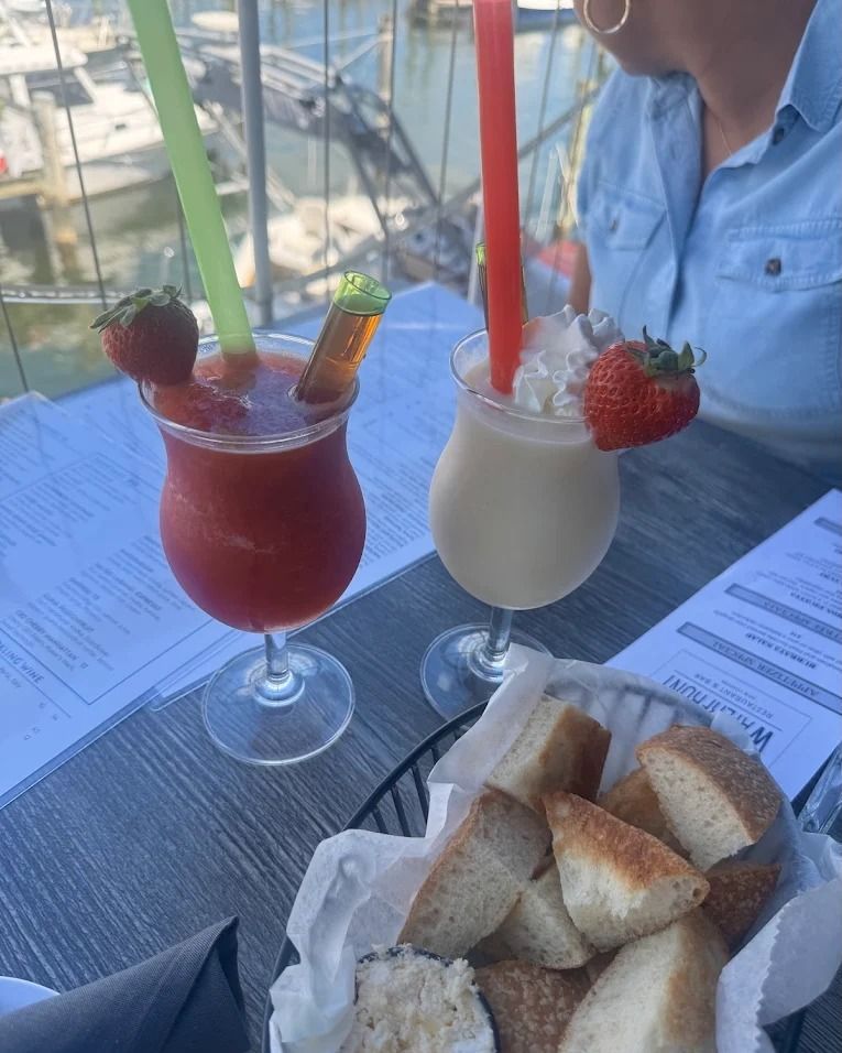 Two cocktails with straws and strawberries, basket of bread, person, outdoor table.