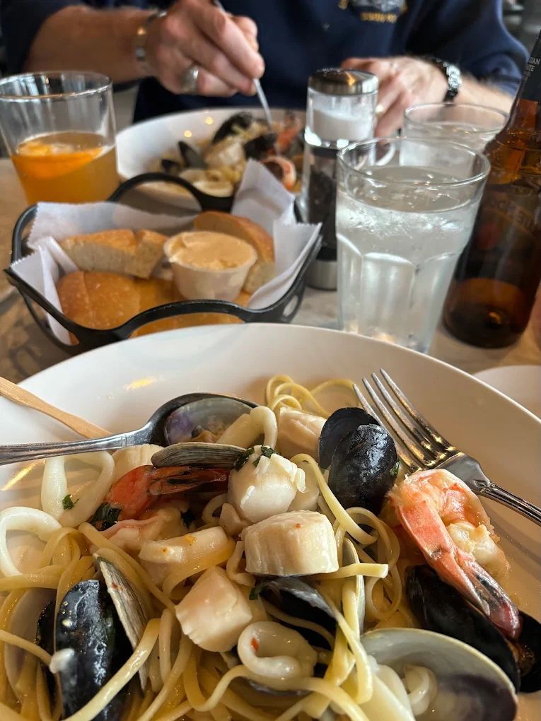 Seafood pasta dish with bread, drinks, and a person eating at a restaurant table.