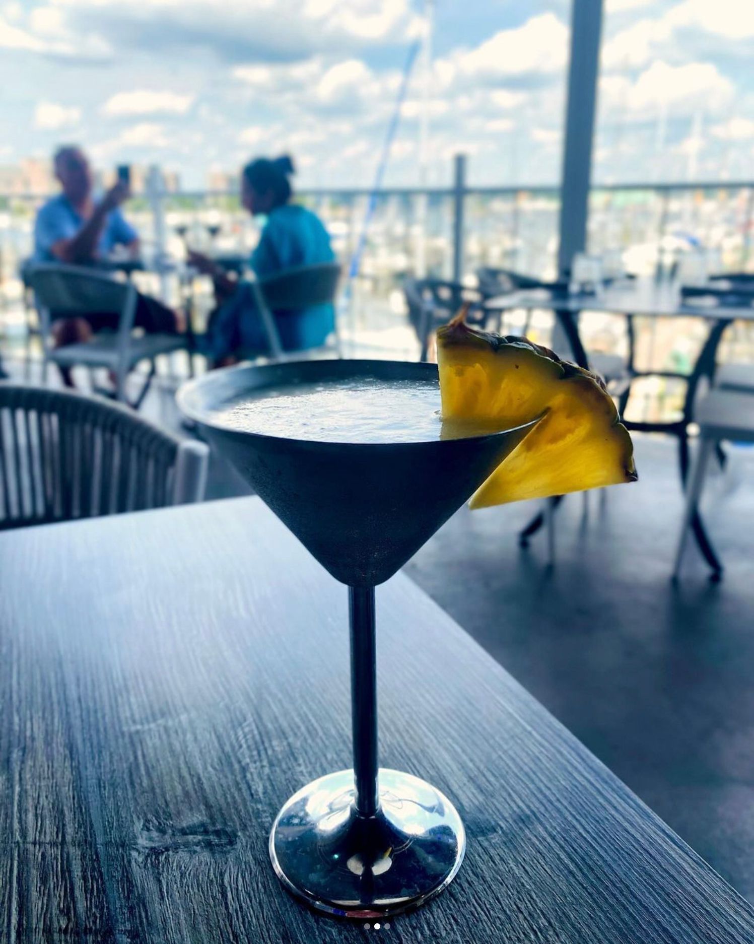Cocktail in a metal martini glass with pineapple garnish on a wooden table, outdoor restaurant setting.