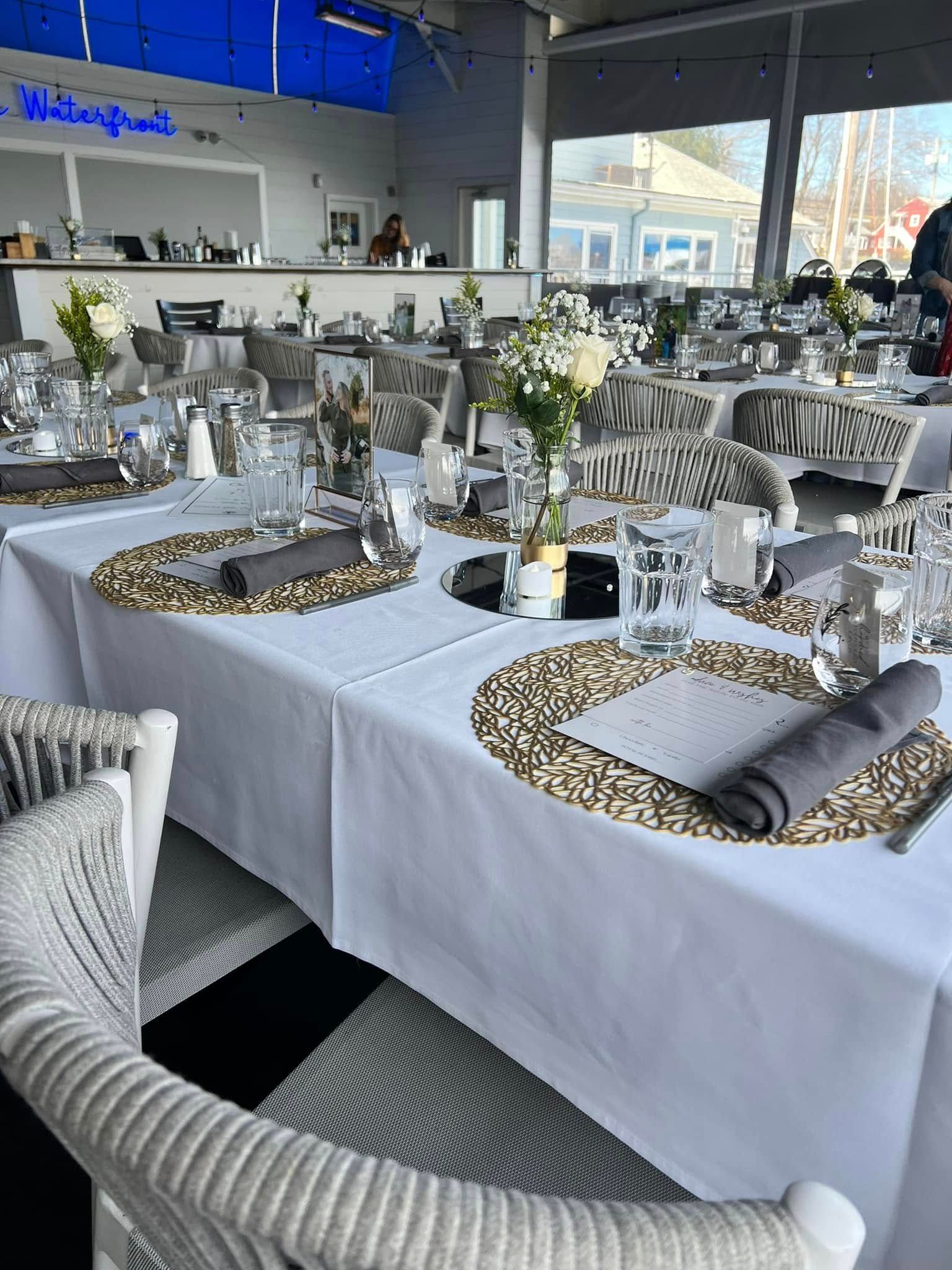 Restaurant tables set for a gathering with white tablecloths, gold chargers, and floral centerpieces.