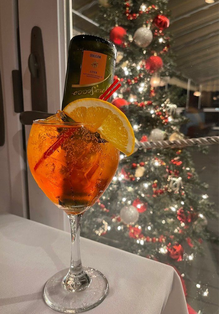 An orange Aperol spritz in a stemmed glass with an orange slice garnish and a bottle of Prosecco, Christmas tree in background.