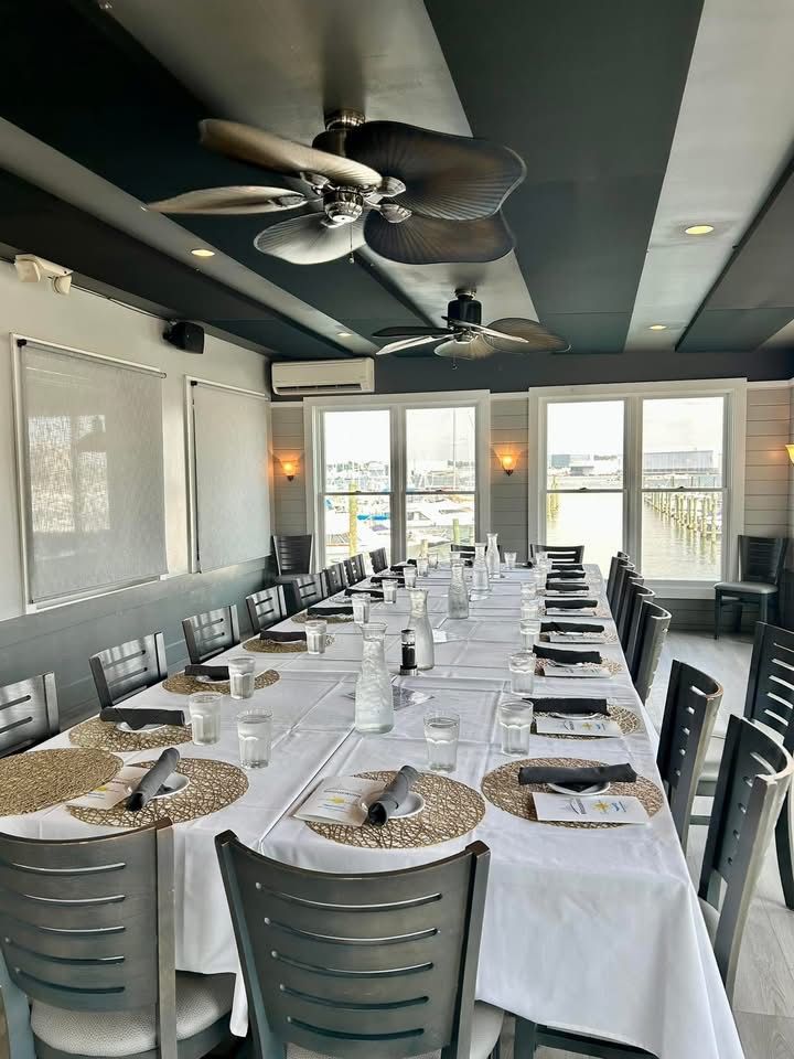 Long dining table set for a meal in a restaurant with large windows overlooking a harbor.
