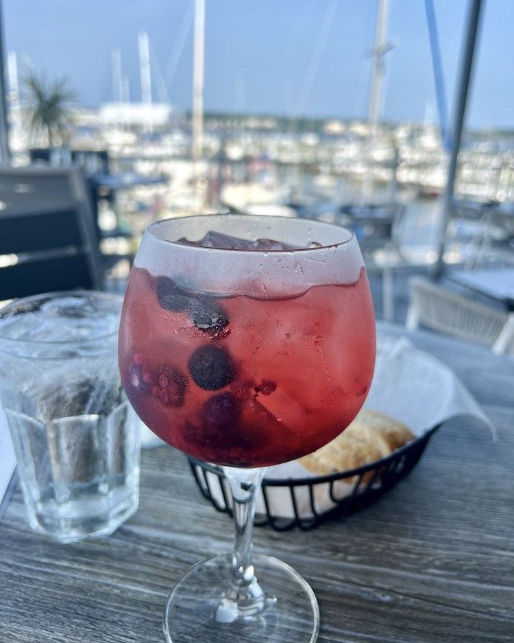 Red cocktail with ice and blueberries in a glass, with a harbor backdrop.