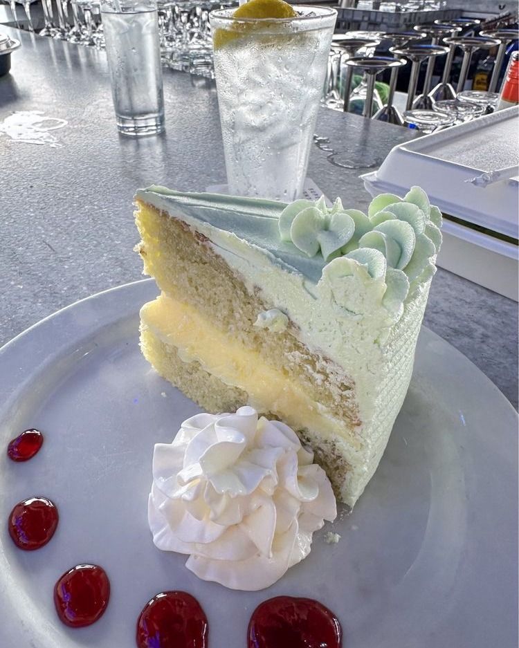 Slice of cake with light blue frosting and whipped cream on a plate, next to a drink.