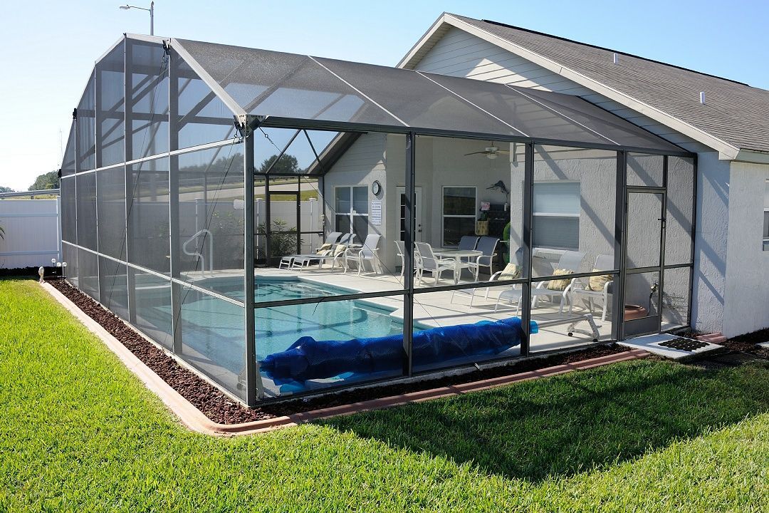 Mesh-covered backyard pool enclosure beside a white house with a blue pool inside.