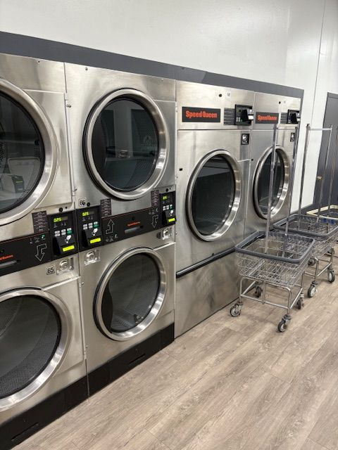 Row of stainless steel commercial laundry dryers and washers in a laundromat setting.